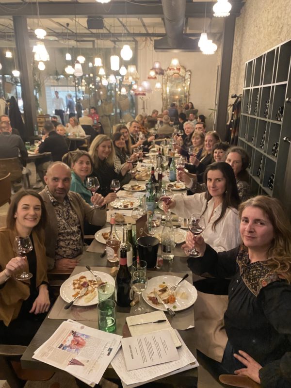 Une longue table rectangulaire et 14 personnes en train de lever leur verre de vin dans un restaurant avec de nombreuses lumières qui pendent du plafond