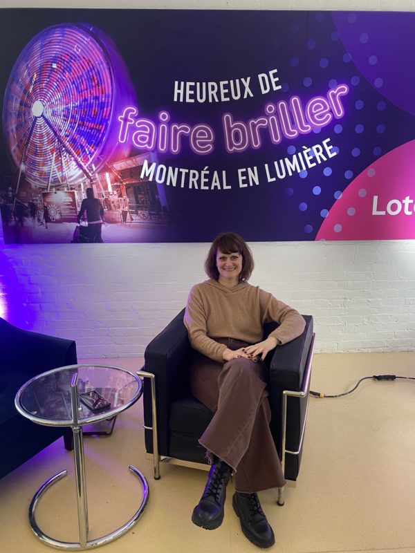 Dame assise dans un fauteuil devant une affiche sur lequel est écrit: Heureux de faire briller Montréal en lumière
