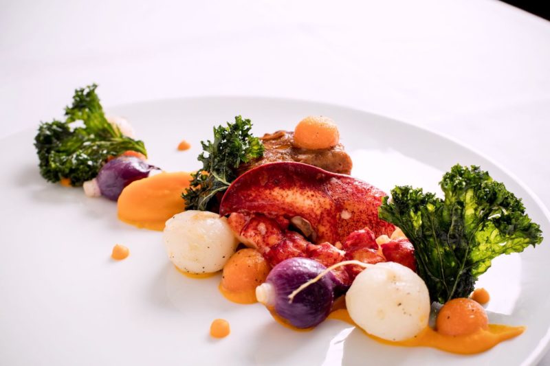 Dans une assiette blanche une pince de homard et quelques petits légumes installés en ligne navet, carotte, herbes
