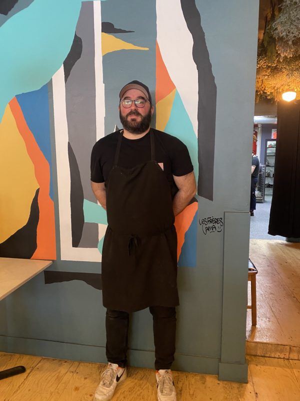 Un chef en noir barbue avec des lunette appuyé contre le mur peint de son restaurant 