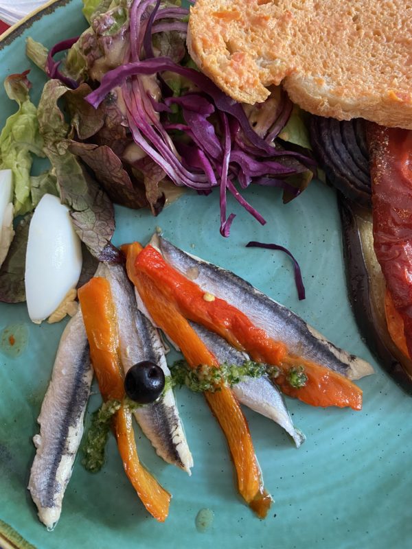 assiette bleue avec des anchois recouvert de poivrons grillés une olive et à côté du pain recouvert de purée de tomate 