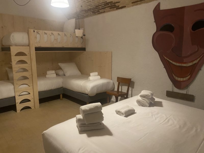 Une chambre avec un grand lit blanc et au fond deux lit superposés  en bois avec une échelle . Au mur une peinture représentant une sorte de diable marron 