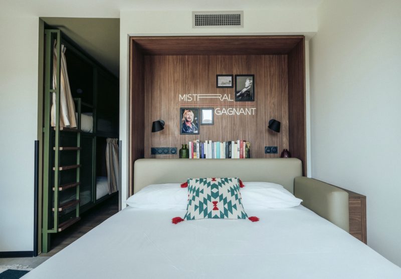 Un lit de face blanc avec un coussin vert et rouge . Ce dernier est encadré dans une alcôve en bois . Sur le côté on aperçoit des lits superposés intégrés dans une alcôve en métal peint vert clair