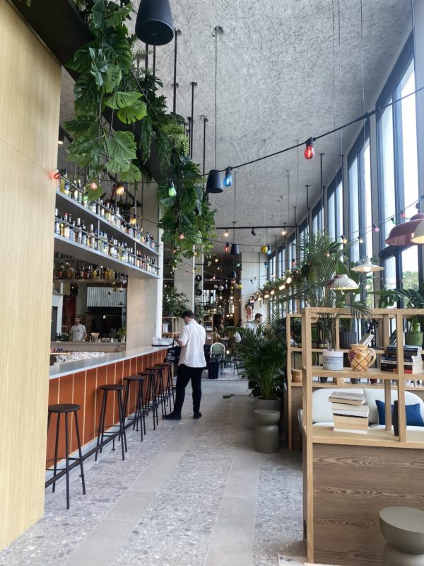 Une enfilade de tables dressées sur la droite de l'image accolées à de grandes baies vitrées et sur la gauche le bar qui se finit au plafond par des plantes vertes